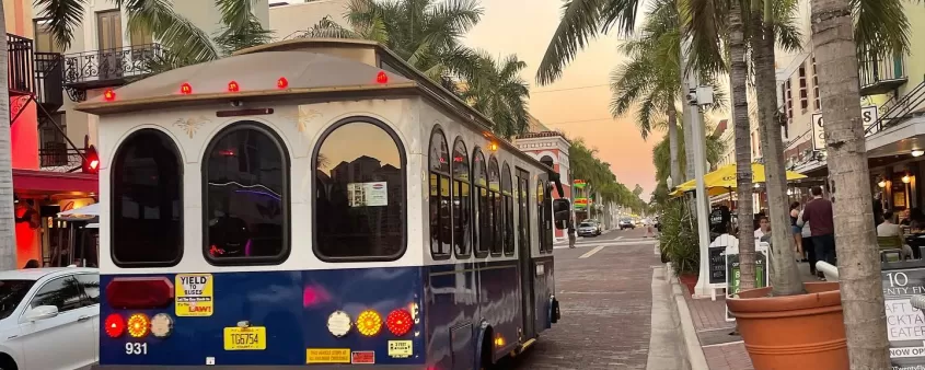 Blue and white trolley driving down a palm-lined brick street at sunset with outdoor diners nearby.